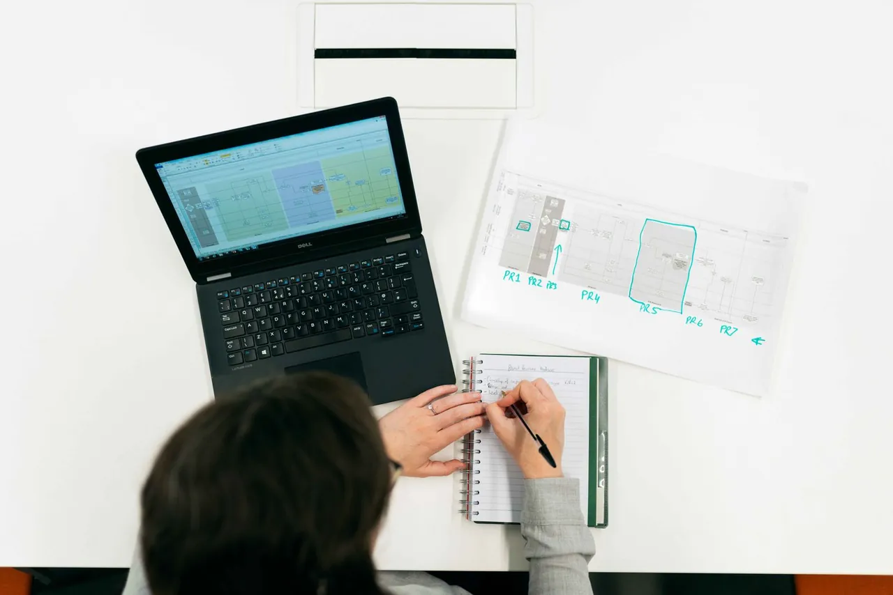 Eine Person macht Notizen neben einem Laptop und einem technischen Plan.