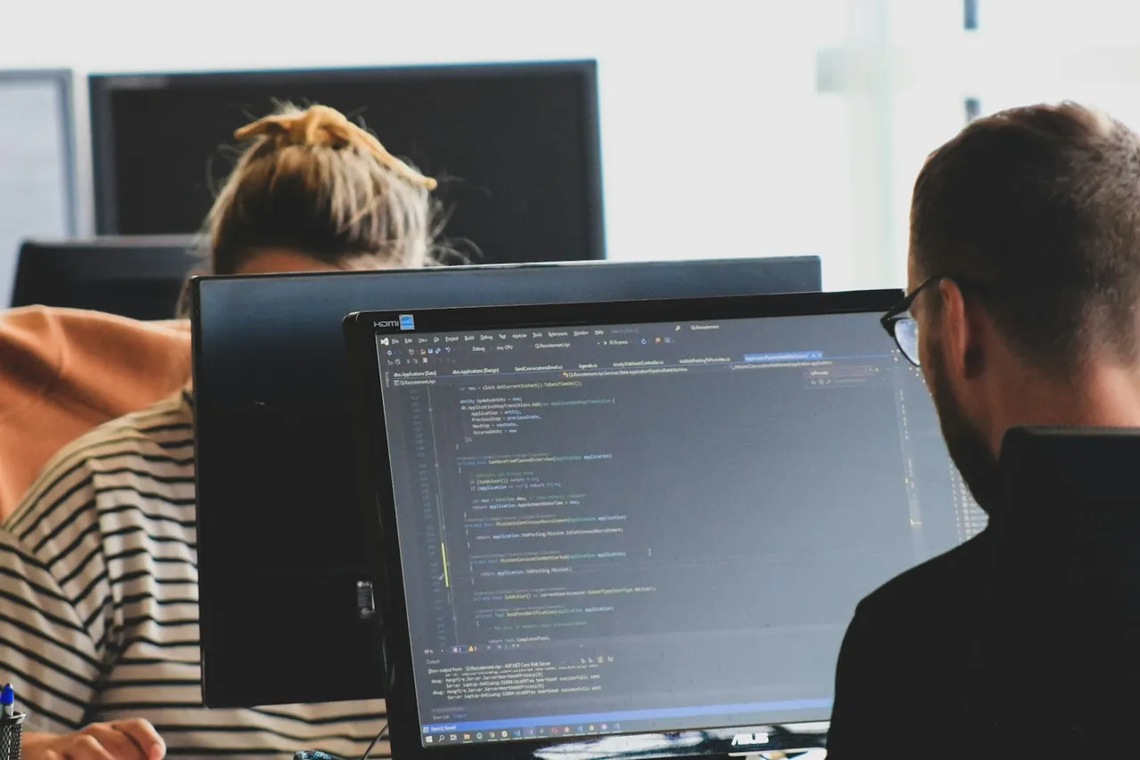 Zwei Personen arbeiten an Bildschirmen mit geöffnetem Programmcode.